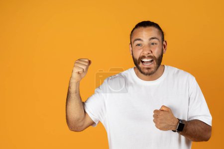 Foto de Feliz millennial europeo hombre con barba en camiseta blanca disfrutar de buenas noticias, victoria, éxito celebra con el puño en alto, grito, aislado en fondo de estudio naranja. Ganar y estilo de vida emociones - Imagen libre de derechos