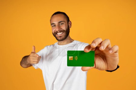 Foto de Positivo millennial europeo hombre con barba en camiseta blanca muestran tarjeta de crédito y el pulgar hacia arriba gesto, aislado en fondo de estudio naranja. Financiación de la recomendación, beneficio de aprobación, ahorro - Imagen libre de derechos