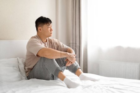 Photo for Lonely pensive upset handsome asian middle aged man wearing pajamas sitting on bed at home, looking through window, suffering from depression, feeling down, copy space, side view - Royalty Free Image