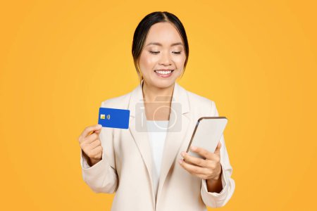 Foto de Mujer milenaria asiática feliz en traje de negocios, tiene tarjeta de crédito, uso de teléfono inteligente para las compras en línea, aislado en fondo naranja. Finanzas bancarias, pagar por la orden, comprar aplicación - Imagen libre de derechos