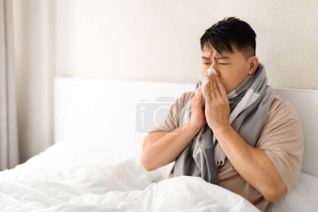 Téléchargez les photos : Gros plan de l'homme asiatique malade soufflant nez liquide dans la serviette tout en étant assis dans le lit à la maison, se sentant mal à l'aise tout en se reposant dans la chambre, ayant la grippe saisonnière ou la fièvre, Copiez l'espace - en image libre de droit