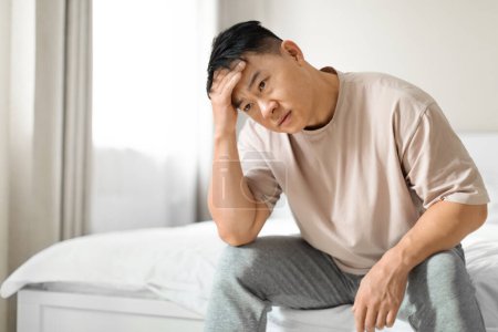 Photo for Depressed middle aged handsome chinese man wearing pajamas sitting on bed alone at home, touching his head, suffering from loneliness or anxiety, copy space. Depression, mental disorders - Royalty Free Image