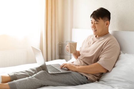 Photo for Relaxed cheerful happy attractive middle aged asian man lying on bed at home, using modern pc laptop, drinking coffee, typing on computer keyboard and smiling, choosing movie, copy space - Royalty Free Image