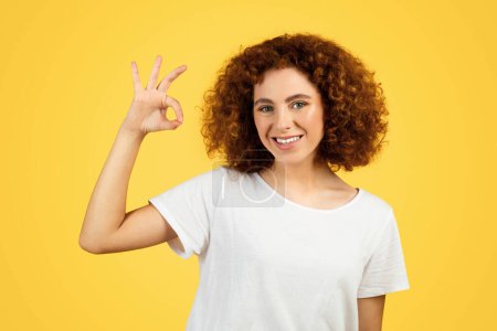 Téléchargez les photos : Souriant jeune étudiante frisée en t-shirt blanc montre ok signe avec la main, isolé sur fond jaune, studio. Les gens positifs émotions, approuver geste, recommandation annonce et offre - en image libre de droit