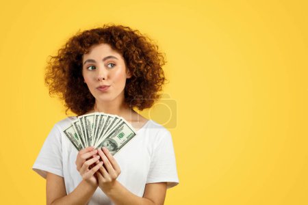 Photo for Smiling pensive funny european young lady, hold money, think, look at copy space, isolated on yellow background. Recommendation financial profit and dream, credit, ad and offer - Royalty Free Image