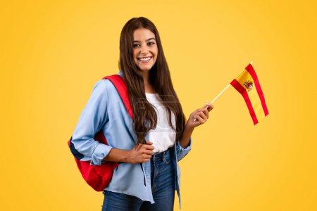 Photo for Happy european young student lady, with Spain flag, combines patriotism with education, isolated on yellow background. Lifestyle, national pride and exchange study, learning Spanish - Royalty Free Image