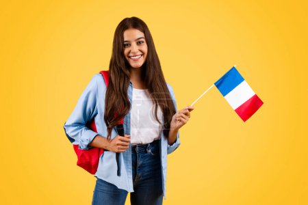 Photo for Cheerful caucasian young student woman, with France flag, combines patriotism with academia, isolated on yellow background. Lifestyle, national pride and exchange study, learning french - Royalty Free Image