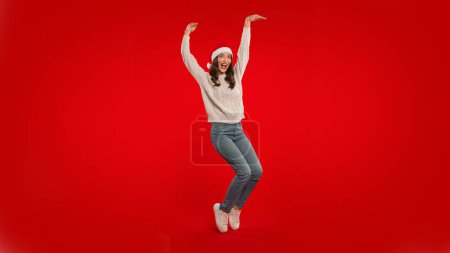 Téléchargez les photos : Joyeux Noël. Funny Young Woman In Santa Hat lever les bras posant sur fond rouge Studio. Femme en jeans décontractés et pull publicité grande offre de Noël. Panorama avec espace vide - en image libre de droit