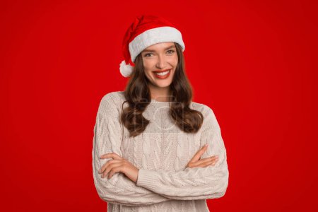 Téléchargez les photos : Jeune femme souriante portant le chapeau de Père Noël et pull décontracté, posant avec les bras croisés sur fond rouge, profitant des vacances d'hiver en studio. Bannière de Noël - en image libre de droit