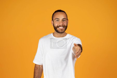 Téléchargez les photos : Confiant homme heureux et sympathique en t-shirt blanc, tendre la main dans un geste accueillant, isolé sur fond orange studio. Partenariat, bonne affaire, réunion et dire bonjour - en image libre de droit