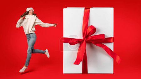 Photo for Energetic woman in winter clothes joyfully jumping beside a giant gift with ribbon bow on red background in studio. Festive excitement and holiday surprises offers. Collage, panorama - Royalty Free Image