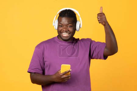 Photo for I Like This Song. Joyful Black Guy Wearing Headphones Listening To Music Playlist Via Phone, Showing Thumbs Up, Approving Musical App Over Yellow Background. Studio Shot - Royalty Free Image