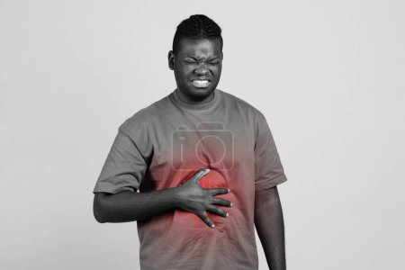 Photo for African american man having stomachache touching aching inflamed abdomen with red spot, posing on gray background, looking at camera. Stomach pain concept. Black and white - Royalty Free Image