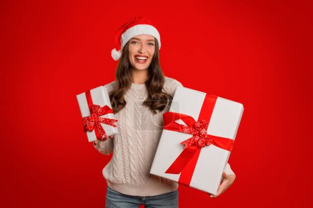 Photo for Cheerful lady holds two beautifully wrapped Xmas boxes against red studio backdrop setting, expressing wonder and charm of Winter holidays season, celebrating joy, smiling to camera - Royalty Free Image