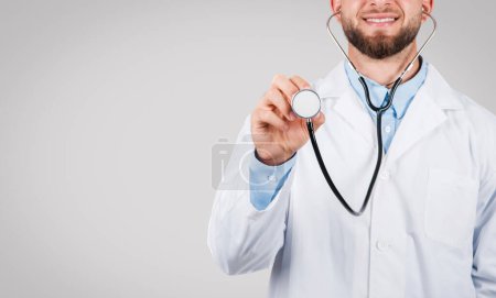 Photo for Confident male doctor with a beard, wearing a white lab coat and blue shirt, smiling while holding a stethoscope towards the viewer, indicating readiness for a medical examination - Royalty Free Image