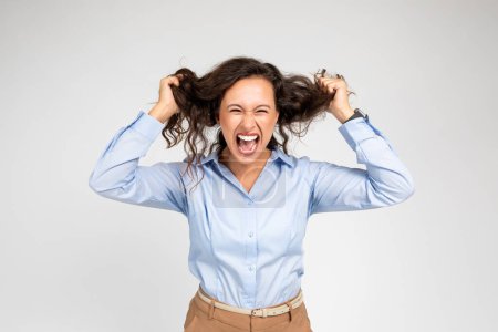 Foto de Mujer europea joven y triste enojada en ropa formal grita, enloquece, arranca el pelo, aislado en el fondo del estudio gris. Problemas de trabajo, estrés, presión y plazo - Imagen libre de derechos