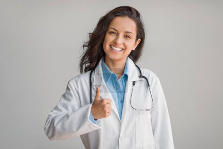 Photo for Happy female doctor in white coat, confidently giving thumbs up gesture, showcasing approval and positivity with a bright smile over light background - Royalty Free Image