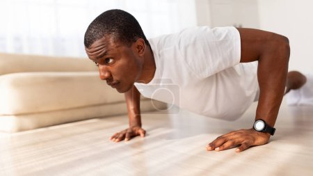 Photo for Motivated athletic young black man wearing white sportswear exercising in his house gym, doing push ups, side view, panorama with free space. Sport at home - Royalty Free Image