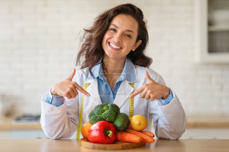 Téléchargez les photos : Joyeuse nutritionniste en blouse de laboratoire avec ruban à mesurer drapé autour de son cou, pointant avec enthousiasme la planche de bois empilée de fruits et légumes vibrants à l'intérieur - en image libre de droit