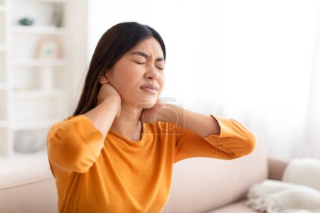 Photo for Unhappy upset young korean woman with long hair sitting on couch at home, touching neck, have strong pain, suffering from osteochondrosis, muscle strain, office syndrome, copy space - Royalty Free Image