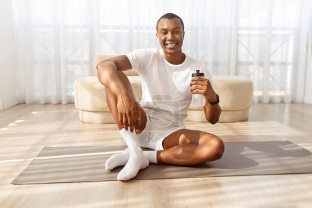 Photo for Positive young african american sporty guy drinking water while having workout at home, sitting on fitness mat, holding bottle and smiling at camera, living room interior, copy space - Royalty Free Image