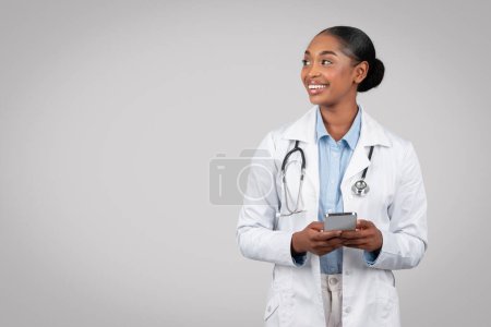 Téléchargez les photos : Joyeux médecin afro-américain confiant en manteau blanc, tapant sur smartphone, regardez l'espace de copie, studio d'arrière-plan gris isolé. Aide médicale à la clinique, travail, app soins de santé - en image libre de droit