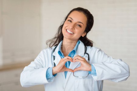 Téléchargez les photos : Portrait de médecin brune joyeuse en manteau blanc et chemise bleue, souriant brillamment et formant la forme du cœur avec ses mains, mettant en valeur l'amour et les soins dans la profession médicale. - en image libre de droit