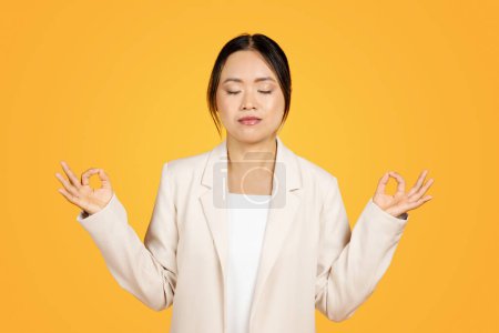 Photo for Young Asian woman in studio work environment engages in meditation, finding moment of peace and calm amid hustle. Mindfulness and mental well-being, zen and breathing exercises - Royalty Free Image