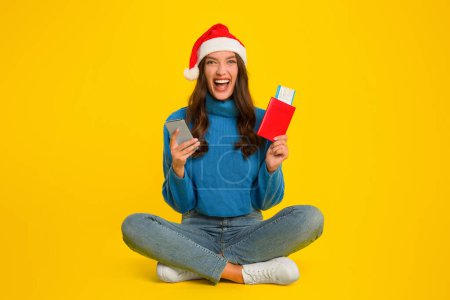 Photo for Joyful young lady in santa hat holding smartphone and passport with boarding pass tickets, booking flight on Christmas, sitting on yellow studio background. Xmas season, winter holiday trip - Royalty Free Image