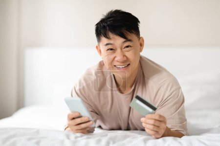 Téléchargez les photos : Heureux homme asiatique mature utilisant Mobile Banking Holding Smartphone et carte de crédit couché sur le lit à la maison le week-end. Application d'achat en ligne, concept de commerce électronique. Espace de copie - en image libre de droit