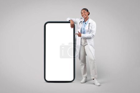 Photo for Positive black millennial doctor in white coat, point finger on big phone with empty screen, isolated on gray studio background. Medical health care recommendation app, website - Royalty Free Image