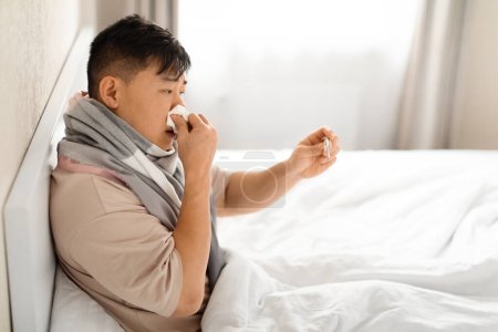 Foto de Hombre asiático de mediana edad enfermo que tiene fiebre, medir la temperatura corporal con termómetro, estornudar en la cama, usar bufanda caliente alrededor del cuello, espacio para copiar, vista lateral - Imagen libre de derechos