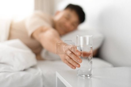 Téléchargez les photos : Homme d'âge moyen asiatique en bonne santé couché au lit à la maison, atteignant avec un verre à main, eau potable après le réveil le matin, espace de copie, fond flou - en image libre de droit