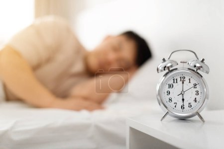 Téléchargez les photos : Concept d'éveil précoce. Homme asiatique dormant par réveil le matin, couché au lit, foyer sélectif sur réveil. Sommeil excessif, il est temps de se réveiller et de se lever. Profondeur faible - en image libre de droit