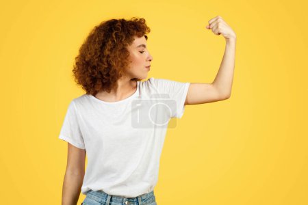 Téléchargez les photos : Force confiante jeune étudiante frisée en t-shirt blanc montre les muscles biceps à portée de main, isolé sur fond jaune, studio. geste de puissance, recommandation annonce et offre - en image libre de droit