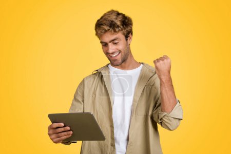 Photo for Stylish young European male confidently holding a digital tablet and making a positive yes gesture, perfectly framed against a clean yellow studio background - Royalty Free Image