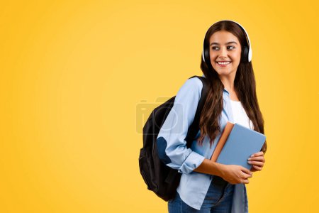 Photo for Smiling calm young student lady typing in headphones, with backpack, enjoy music, lifestyle, look at copy space, isolated on yellow background. App for listen lesson, audio app - Royalty Free Image