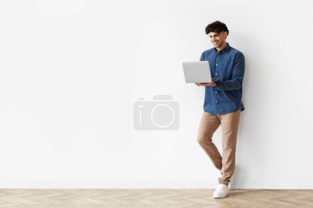 Photo for Full Length Of Smiling Young Arab Guy Using Laptop Leaning On White Studio Wall, Working Online And Websurfing At Home. Blank Space For Online Offer Advertisement - Royalty Free Image