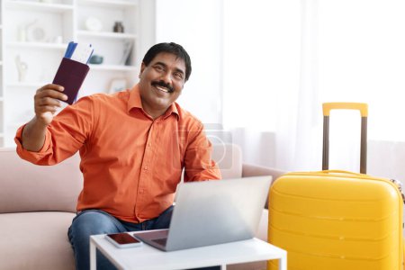 Photo for Online tickets booking. Happy mature indian man using laptop and buying tour at internet travel agency, posing near suitcase at modern home interior, showing tickets. Vacation plans concept - Royalty Free Image