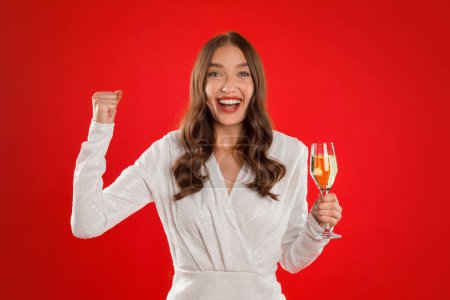 Foto de Holiday Joy. Emocionado joven elegante mujer en vestido blanco Gestos Sí y sosteniendo la copa de vino espumoso sobre fondo de estudio rojo, sonriendo a la cámara celebrando el año nuevo - Imagen libre de derechos