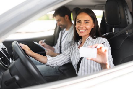 Photo for Successful Driving License Test. Instructor Man Taking Notes While Happy Driver Pretty Young Woman Showing Her Driving License. Lady Driving Sitting In Car. Passed Exam In Auto School Concept - Royalty Free Image