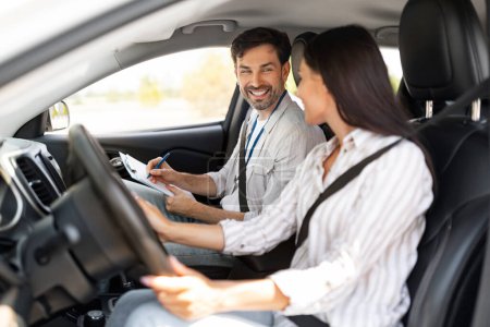 Photo for Handsome man instructor examinating happy lady student, taking notes at chart while sitting by cheerful pretty young woman driving auto, side view, copy space. Driving school concept - Royalty Free Image