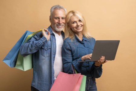 Photo for Cheerful attractive caucasian elderly man and woman shopping on Internet together, using laptop computer pc, holding purchases, beige background. Retail, ecommerce - Royalty Free Image