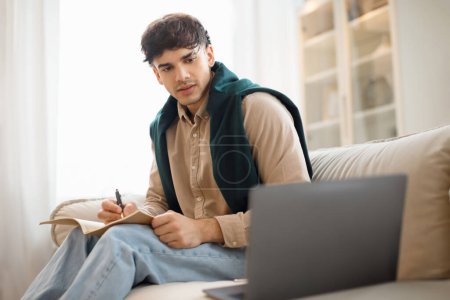 Photo for Distance Education. Young focused Arabic man using pc laptop sitting on sofa, writing in notebook while learning remotely at home. Serious millennial man watching online webinar, taking notes - Royalty Free Image