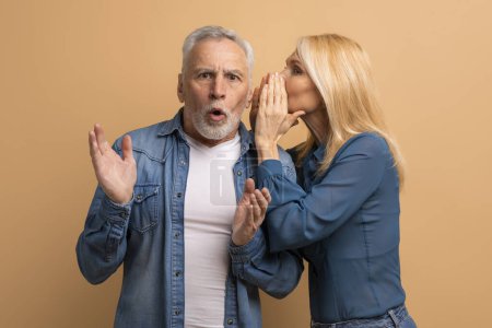 Photo for Attractive mature blonde woman sharing secret or whispering gossips into her husband ear, beige studio background. Loving elderly spouses sharing rumors. Communication in marriage - Royalty Free Image
