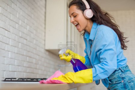Téléchargez les photos : Une jeune femme enthousiaste portant une chemise bleue et des gants jaunes nettoie le comptoir de la cuisine, profitant de sa musique sur des écouteurs roses, chante tout en tenant un vaporisateur et un chiffon - en image libre de droit