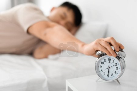 Photo for Morning Awakening. Sleepy Mature Asian Man Hand Turning Off Alarm Clock Waking Up Early Lying In Bed In Bedroom At Home. Time To Wake-Up And Get Up Concept. Shallow Depth - Royalty Free Image