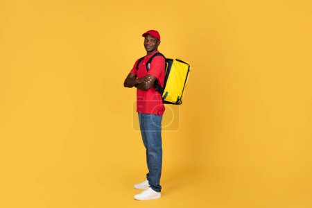 Foto de Hombre seguro positivo en camiseta roja, gorra y jeans con mochila de entrega amarilla. Trabajo profesional de mensajería, buen servicio y estilo de vida, sobre fondo de estudio naranja, longitud completa - Imagen libre de derechos