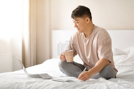 Téléchargez les photos : Portrait d'homme asiatique mature assis dans le lit avec ordinateur portable, Bloc-notes et café, Heureux homme du Moyen Âge utilisant l'ordinateur dans la chambre à coucher pour le travail en ligne et l'éducation, bénéficiant freelance, Copier l'espace - en image libre de droit
