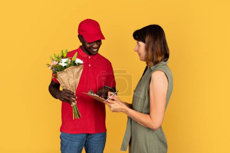 Photo for Smiling millennial black guy courier hold bouquet of flowers, gives clipboard for sign to european lady, isolated on orange studio background. Delivery gift at home, work and service for holiday - Royalty Free Image